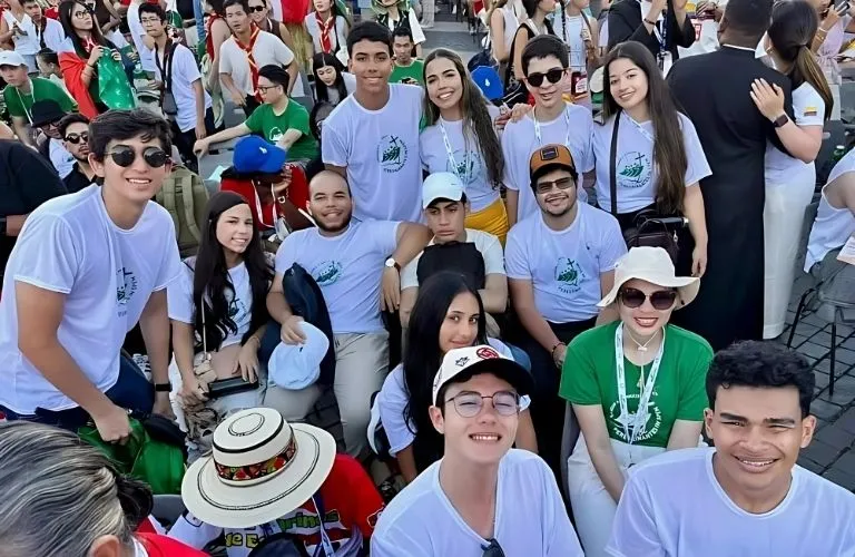Grupo de jovens sorrindo e posando juntos em área externa durante evento, usando camisetas brancas e verdes com o logotipo do Jubileu 2025.